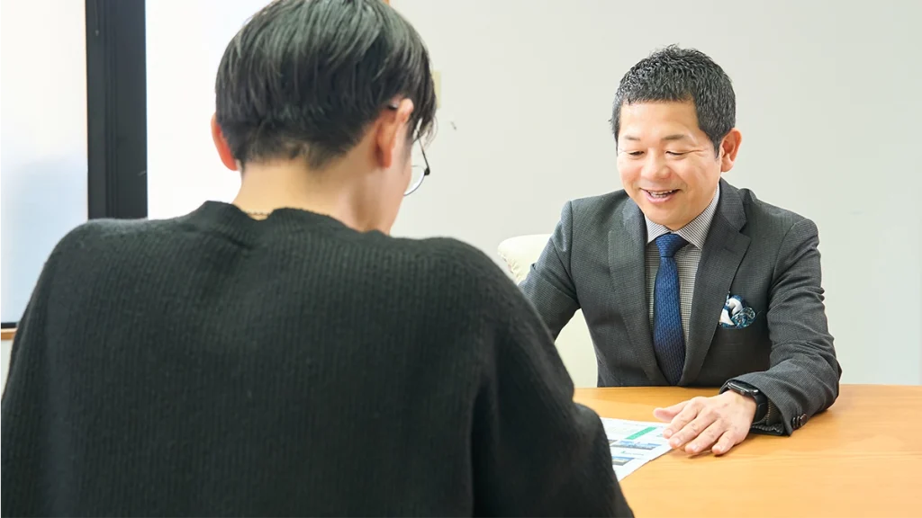 男性スタッフが机に広げた物件資料に手を添え、視線を資料に落として笑顔で説明をしている。